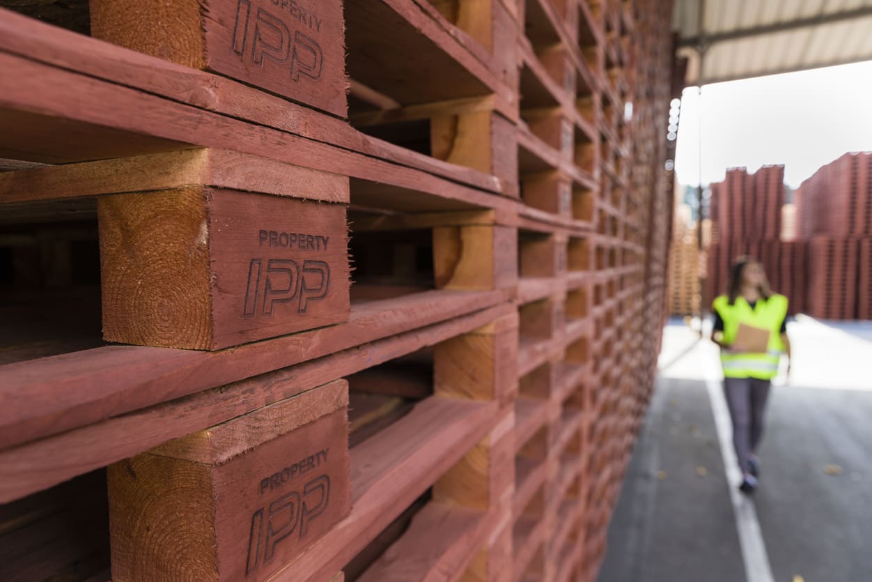 A stack of brown wooden IPP pallets with a women in high vis walking in the background