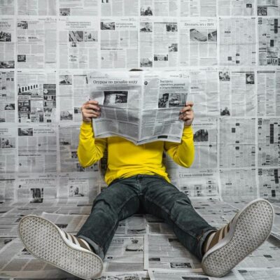 Man wearing yellow top reading a newspaper. The background is made up of newspapers.