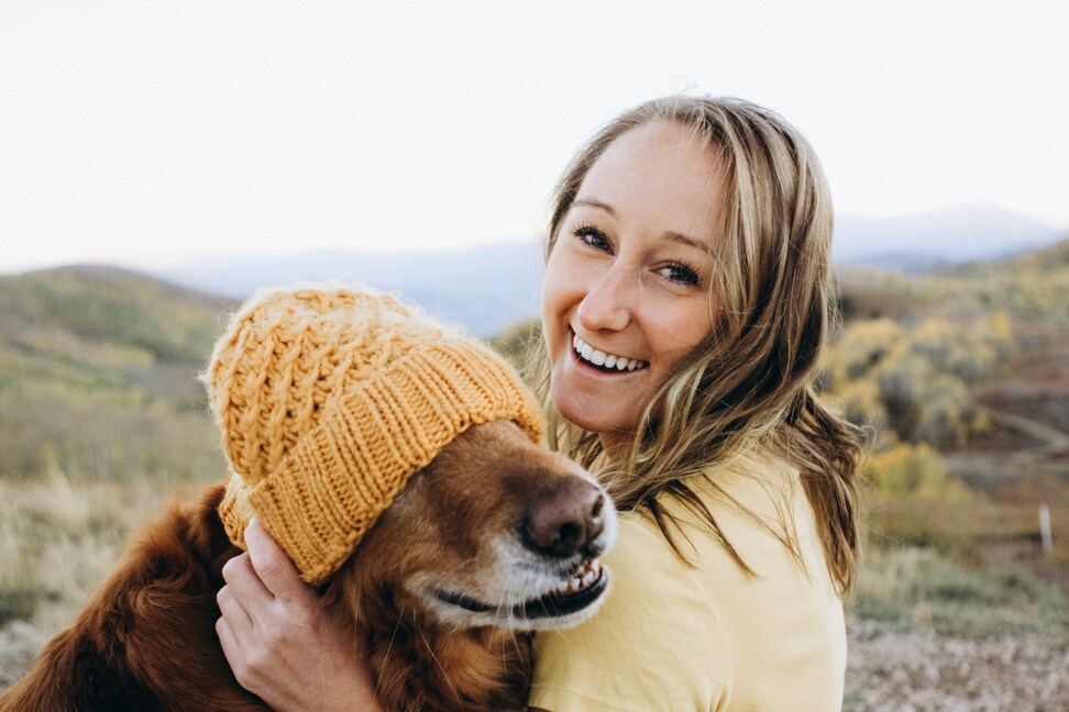 Golden retriever wearing a yellow woolly hat being hugged by a woman