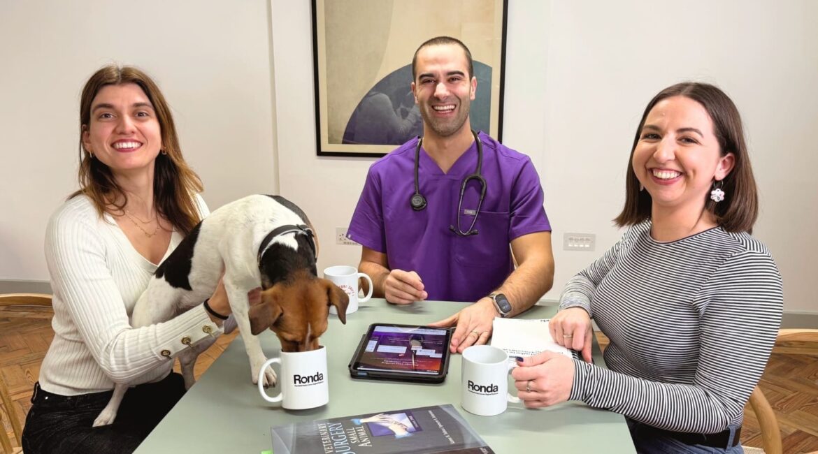 Ronda veterinary team pictured sat round a table. A small terrier has its nose in a mug that is on the table.