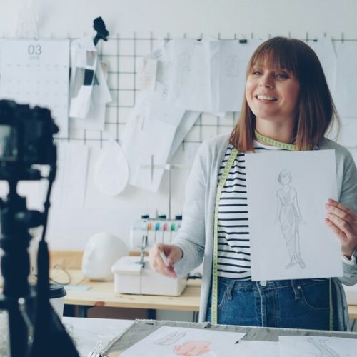 Woman talking into a camera, holding up a sheet of paper as a demonstration in front of a desk